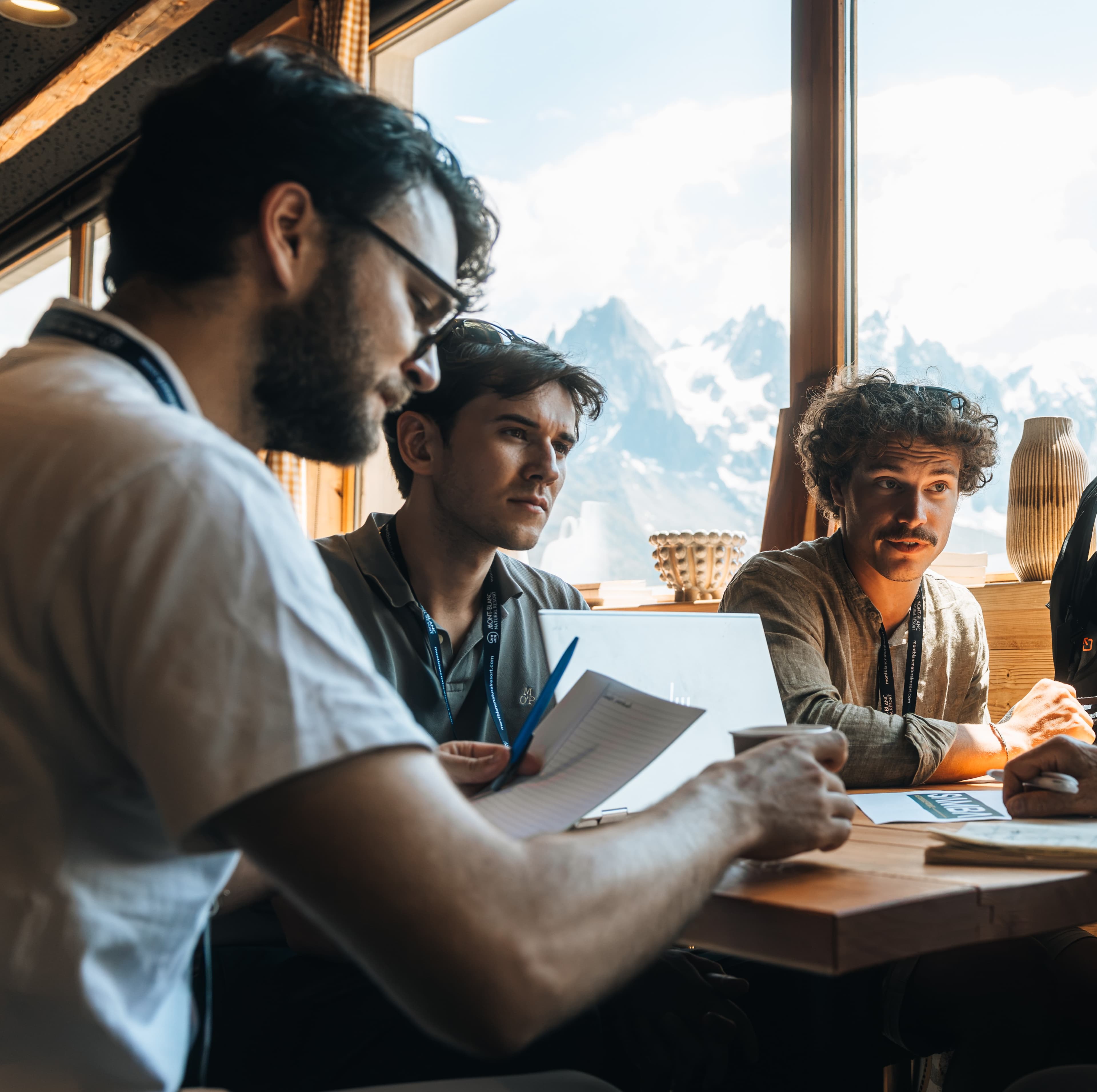 Marcus, Anthony, and Paul reviewing notes together at a table in Chamonix, with the Mont Blanc massif visible through the window behind them