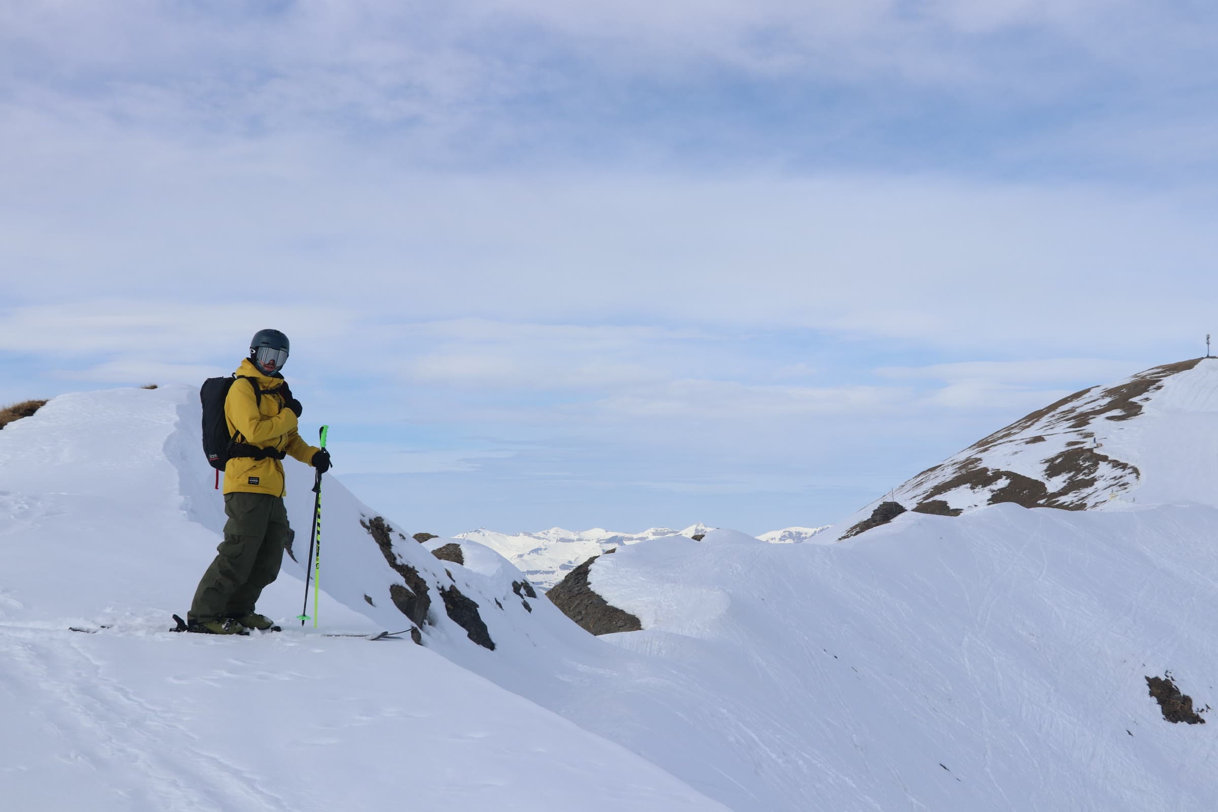 A backcountry skier pausing on a snow ridge, backpack on, looking out over the Alps