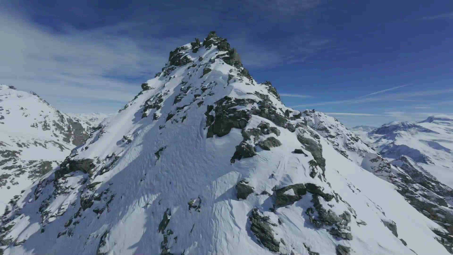 Drone shot flying toward a snow-covered rocky alpine peak under a blue sky